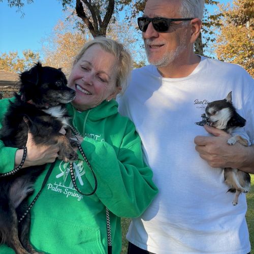 Two smiling adults pose outdoors with two small dogs; the woman in a green hoodie holds a black dog, while the man in a white shirt cradles a lighter-colored dog.