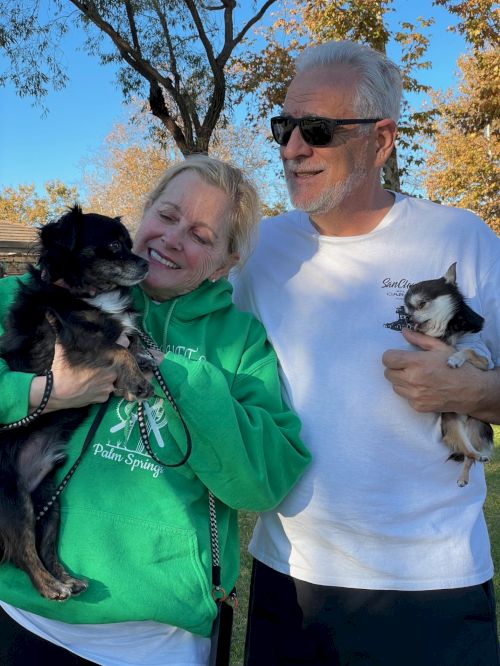 Two smiling adults pose outdoors with two small dogs; the woman in a green hoodie holds a black dog, while the man in a white shirt cradles a lighter-colored dog.