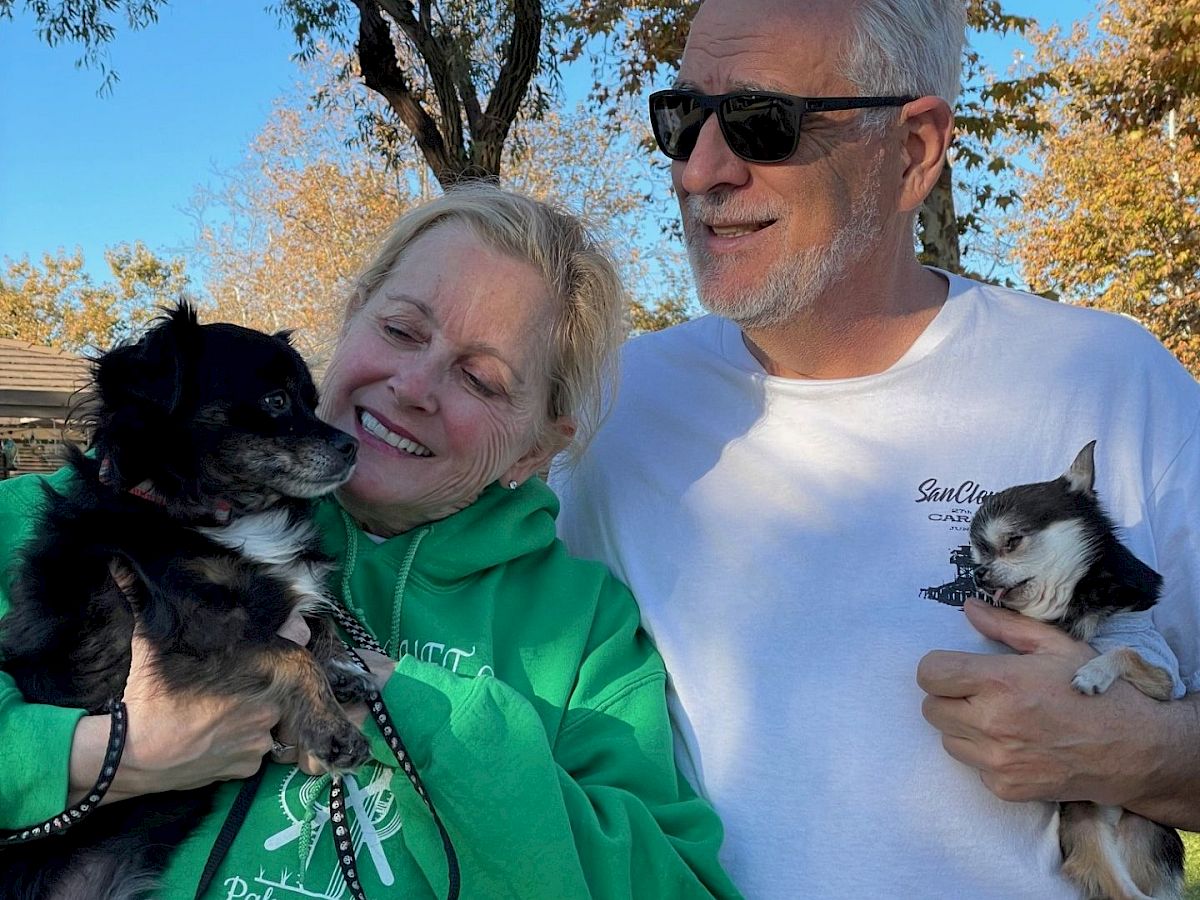 Two smiling adults hold small dogs outdoors on a sunny day, wearing casual clothes; a tree-lined park background.