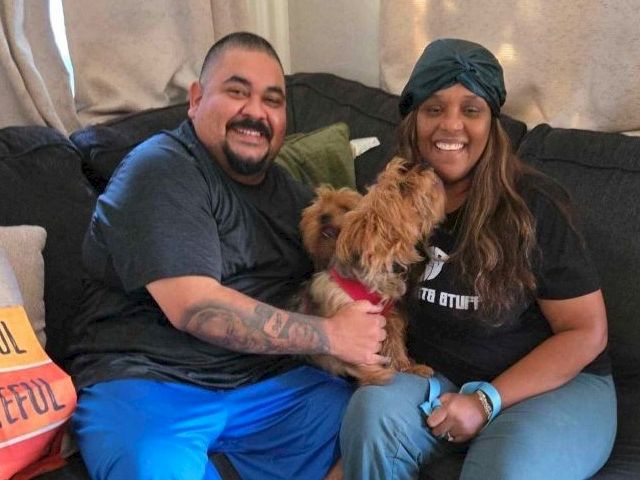 A couple sits on a couch with their small dog between them, smiling warmly for the photo while holding the pup.