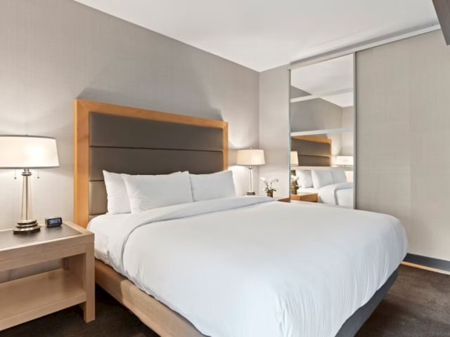 A modern hotel room with a large white bed, wooden headboard, matching nightstands, two lamps, a mirror, and neutral-toned decor. The scene is bright and tidy.