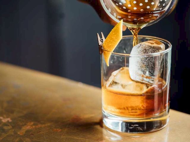 A whiskey glass with ice, a citrus twist garnish, and a gloved hand pouring whiskey over it, on a wooden bar surface.