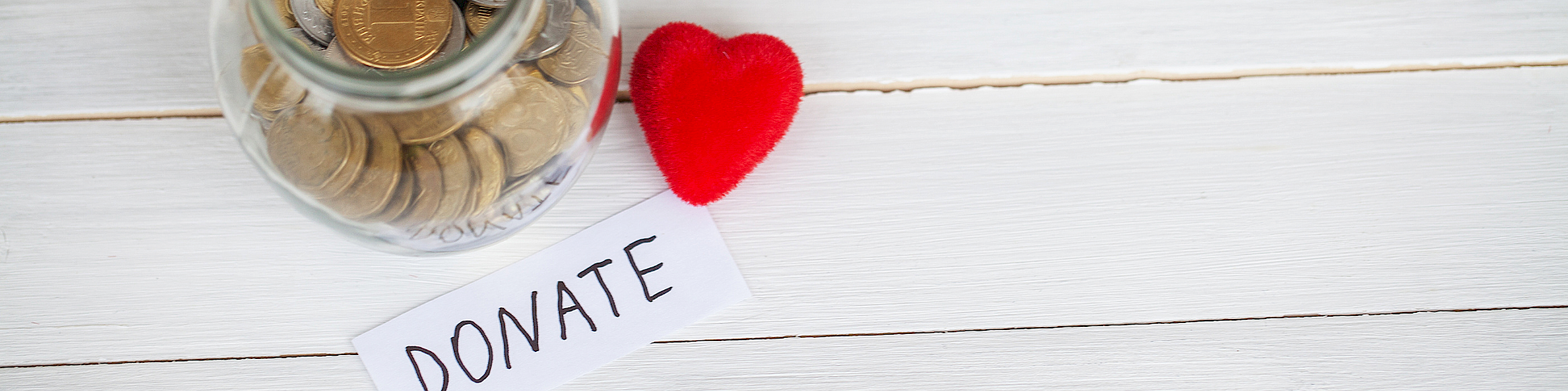 A glass jar filled with coins sits next to a red heart and a note that reads &ldquo;DONATE&rdquo; on a white wooden surface.