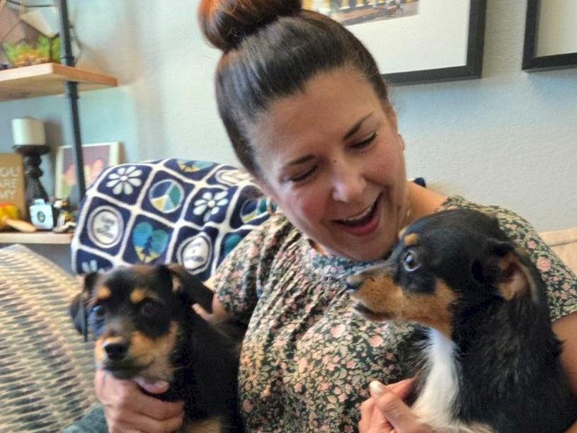 A woman smiles while holding two small dogs, one in each arm, on a cozy sofa with a patterned blanket nearby.
