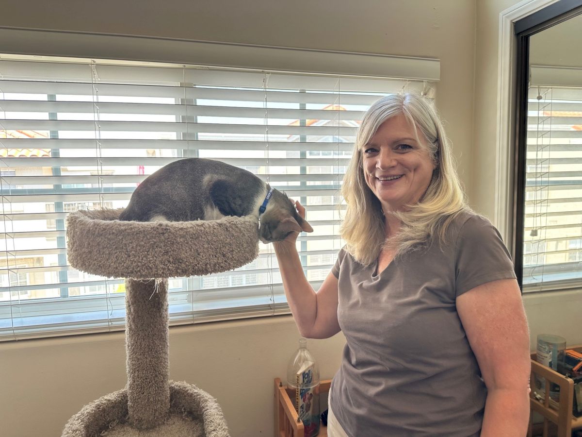 A smiling woman stands next to a cat perched on a tall beige cat tree by a window, both looking content in a bright room.