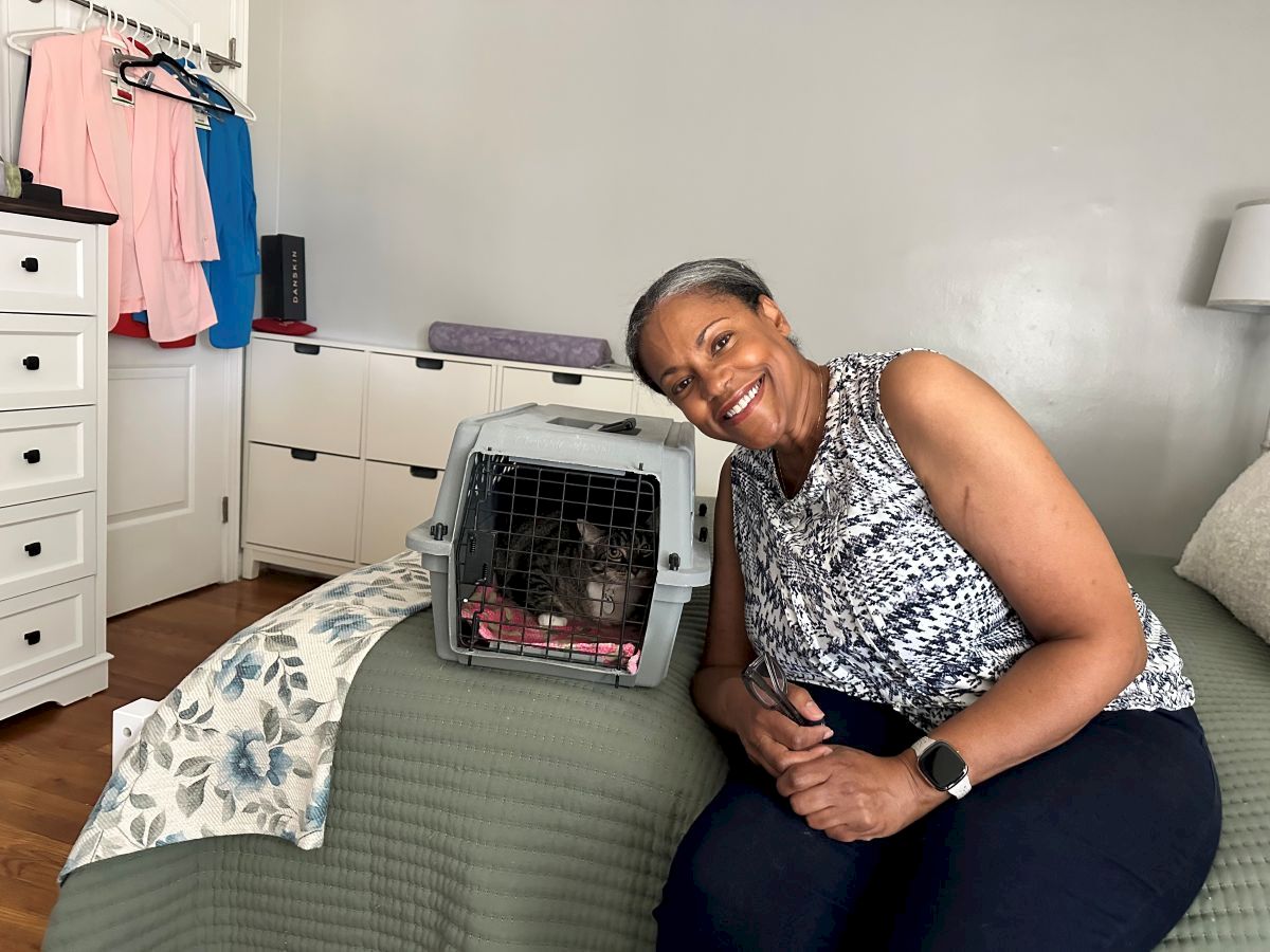 A smiling woman sits on a bed beside a pet crate with a small dog inside, in a tidy bedroom with clothes on a rack in the background.