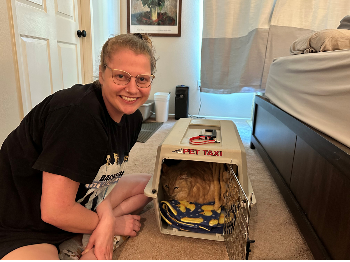 A smiling young person sits on a carpet beside a small dog crate labeled &ldquo;PET TAXI,&rdquo; with a tiny dog inside.