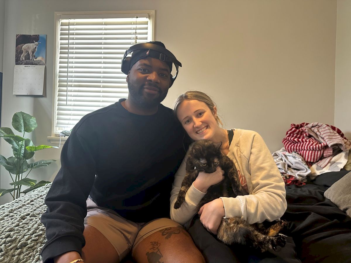 A couple sits on a bed with a small dog, smiling at the camera in a cozy, lived-in room with plants and clutter.