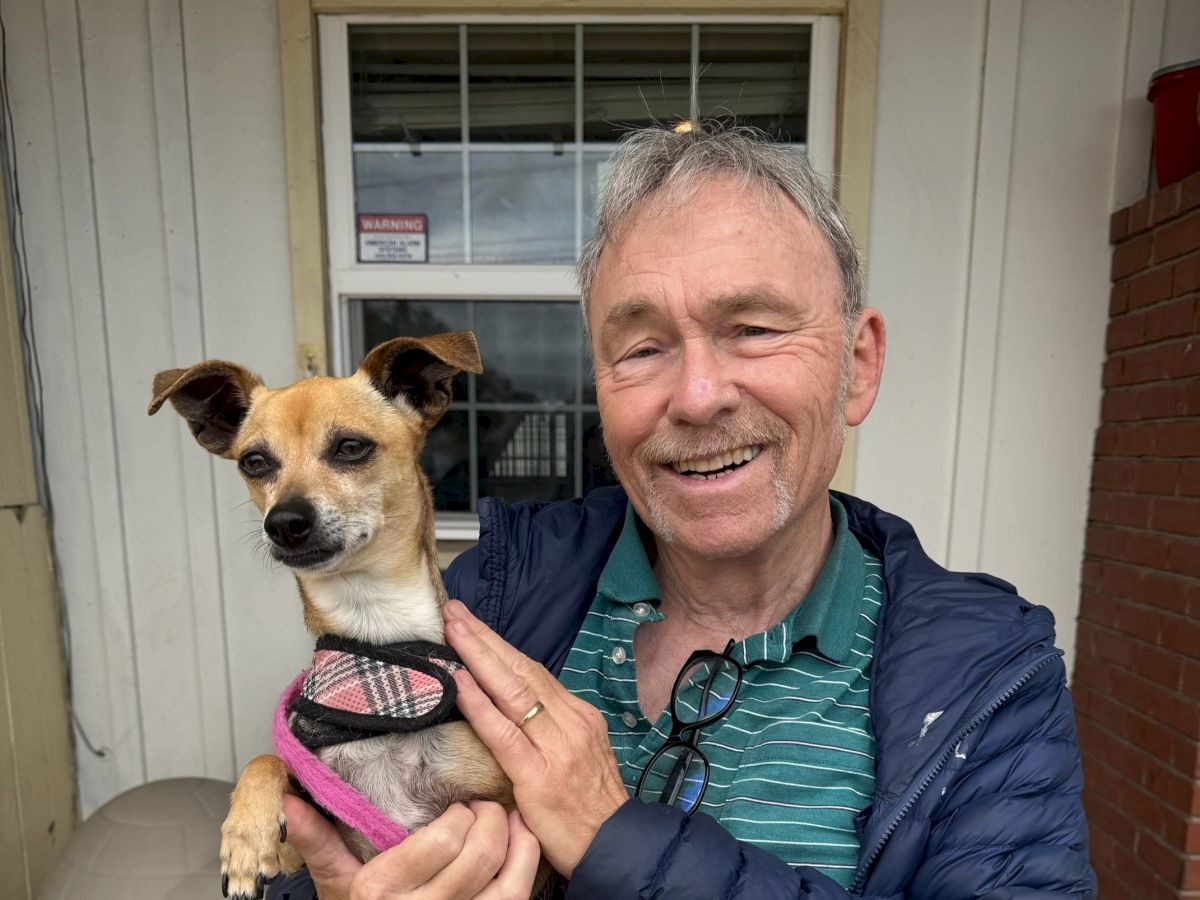 A man in a blue jacket holds a small tan and white dog wearing a pink harness outside a building, both smiling.