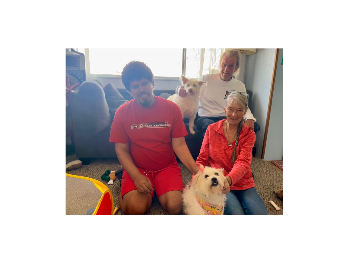 A man in a red shirt sits with two dogs while an older couple smiles behind them in a cozy living room, with one dog wearing a colorful collar.