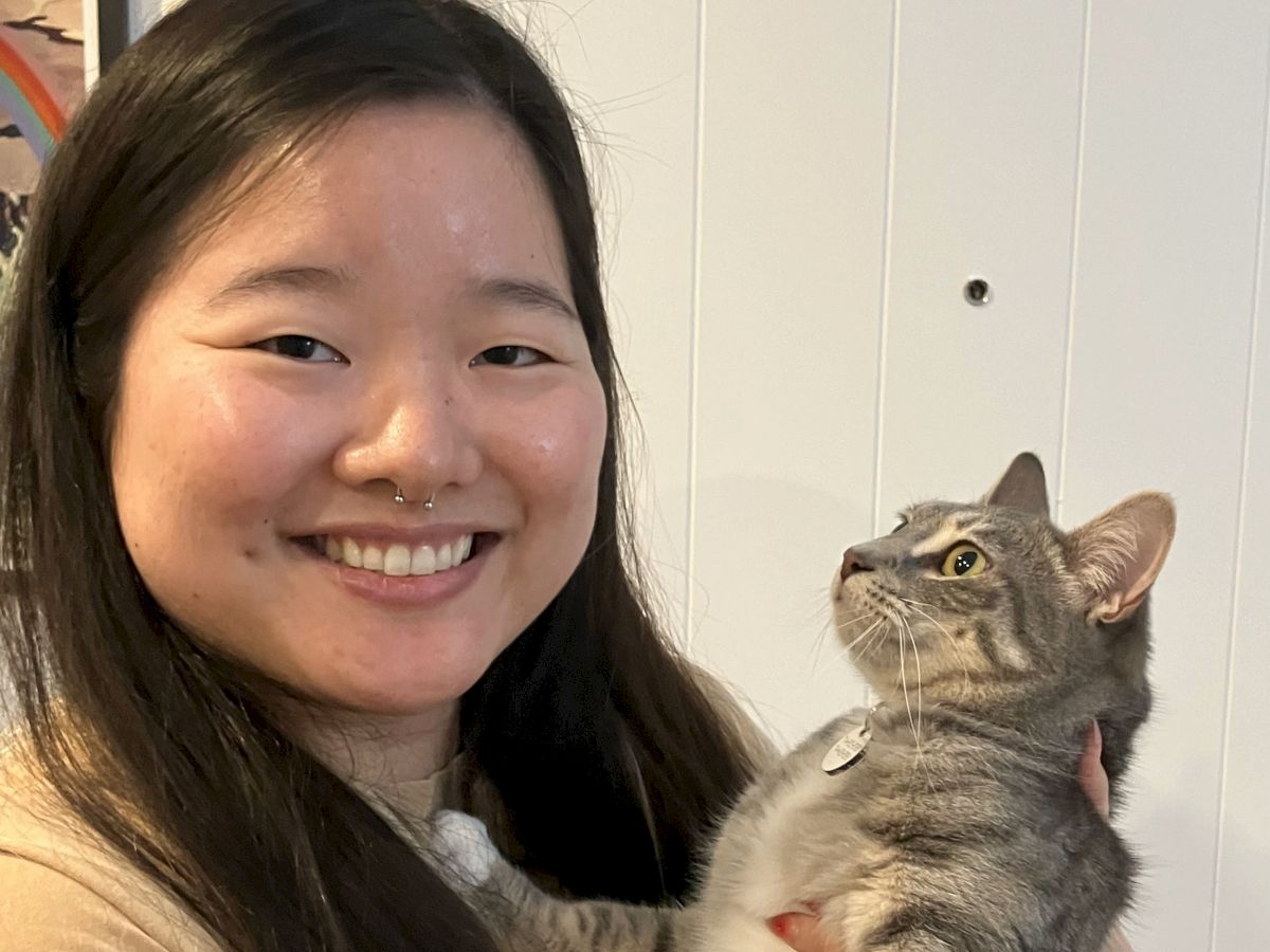 A smiling woman holds a grey tabby kitten indoors, both looking at each other with warmth and affection, bright and cozy vibe.
