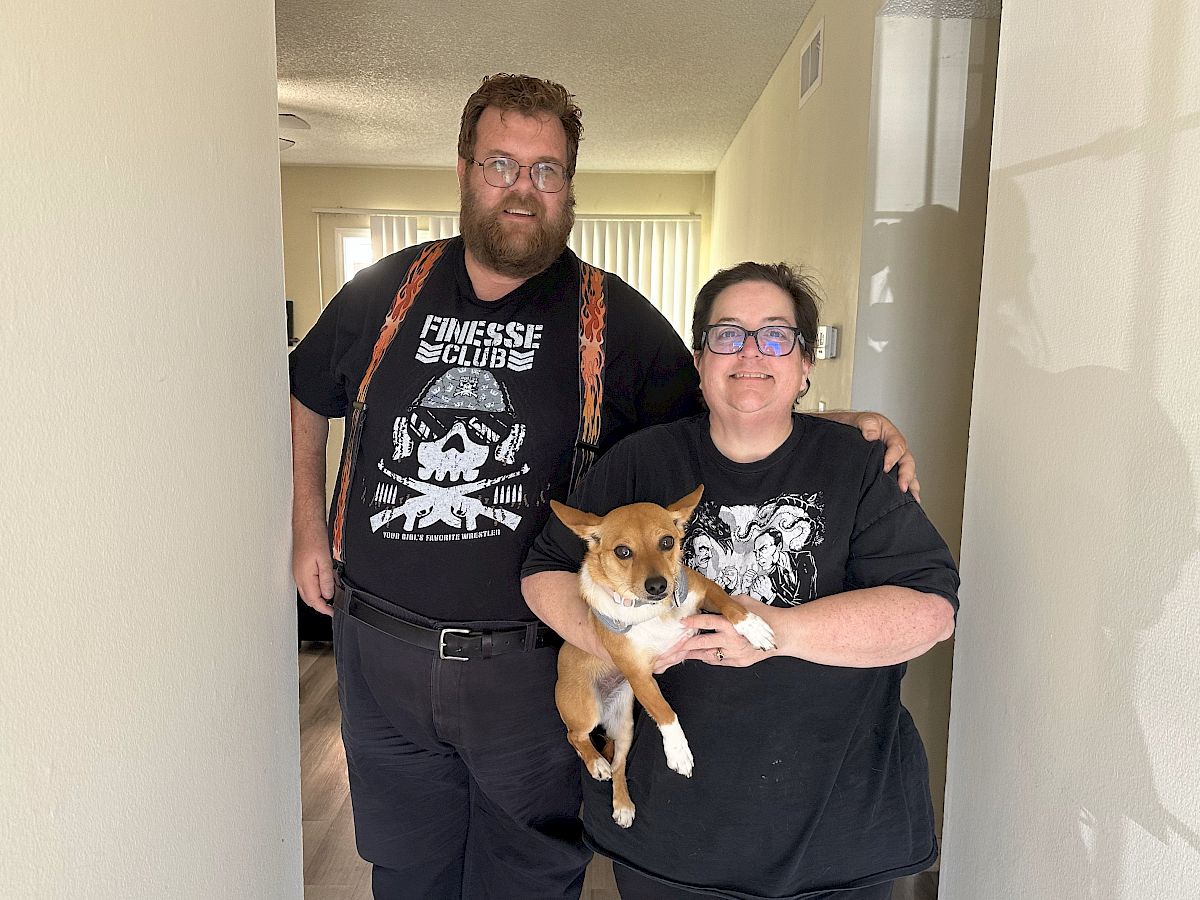 Two people stand in a hallway with a small dog; the man left wears a band tee and suspenders, the woman right holds the dog, all smiling.