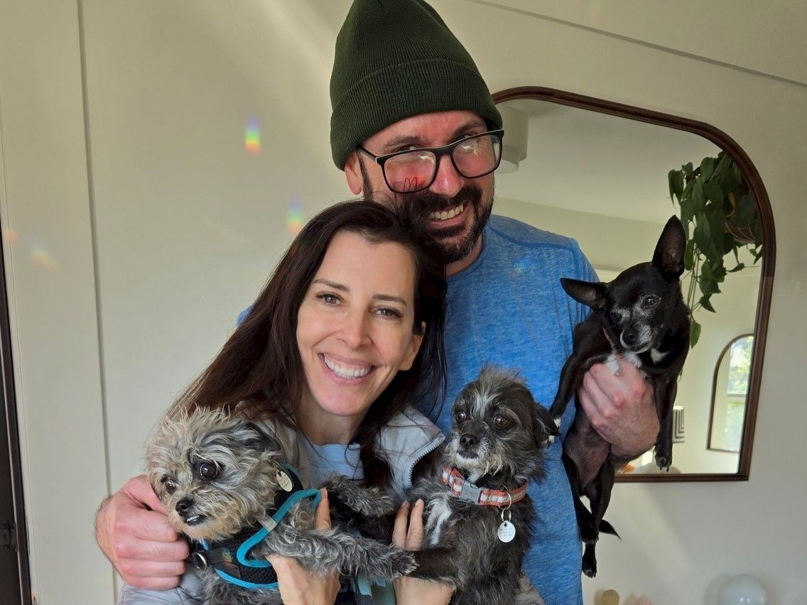 A happy couple holds four small dogs indoors, smiling at the camera, with one man wearing a green beanie and glasses, and a round mirror in the background.