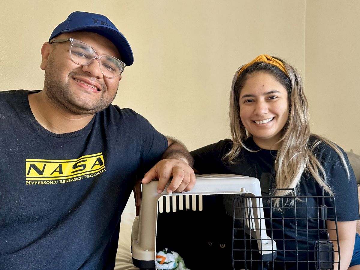 Two people sit smiling beside a pet carrier with a small dog inside, suggesting a pet adoption or transport moment.