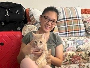 A smiling woman with glasses holds an orange kitten on a floral sofa, with a red suitcase and backpack in the background.