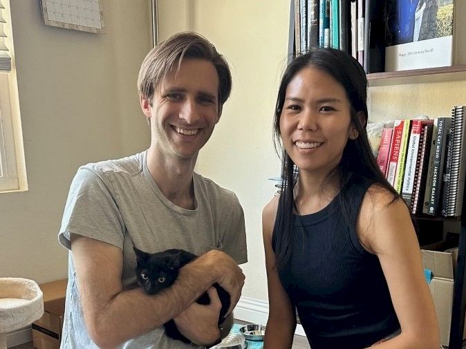 Two people smile in a cozy room; a man holds a black cat, while a woman stands beside them near a bookshelf.