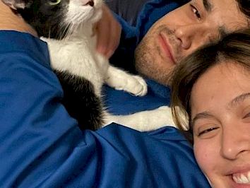 A couple cuddles with a black-and-white cat on a bed, friendliest smiles as they hug the cat close, cozy moment together.