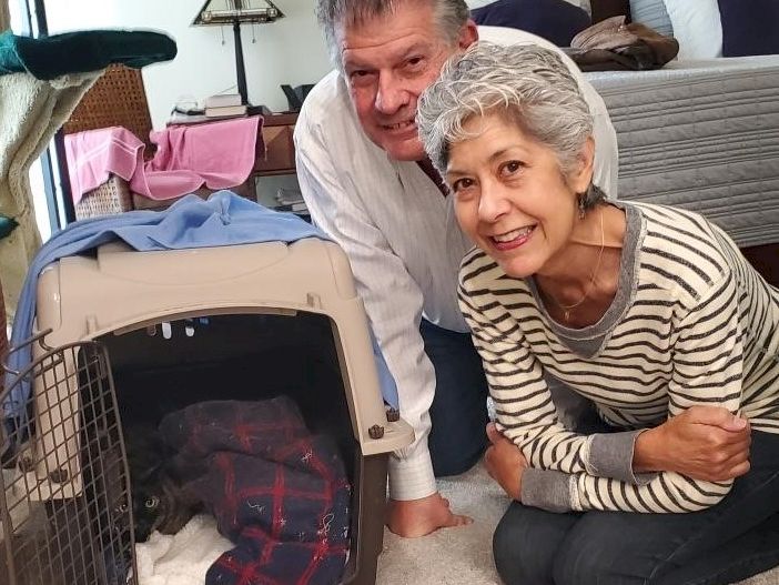 Two smiling seniors pose by an open pet crate with a small dog inside, in a cozy bedroom setup.