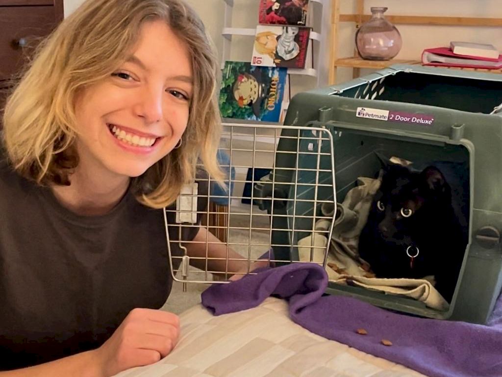 A smiling person sits beside a pet crate with a black cat inside, a purple blanket draped nearby, and cozy home decor in the background.
