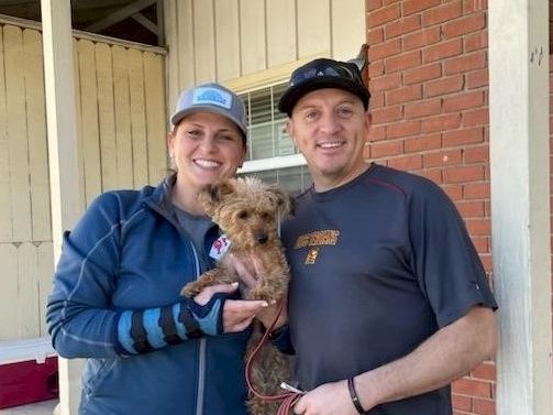 A smiling couple posing with a small fluffy dog outdoors, standing by a brick wall and porch, both wearing caps and casual outdoor attire.