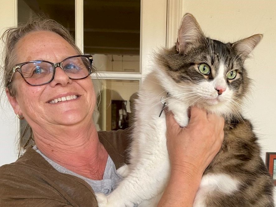 A smiling woman with glasses holds a fluffy gray-and-white cat by the chest, both looking off to the side, indoors.