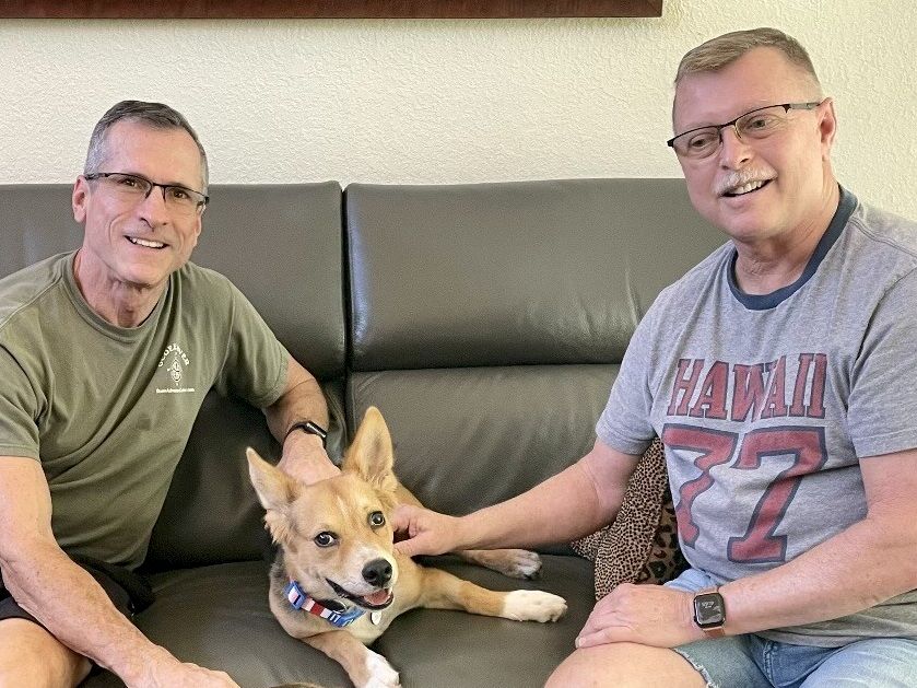 Two smiling men sit on a couch with a happy dog between them, all relaxed and posing for a casual photo.