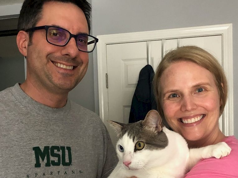 A smiling couple holds a white and gray cat indoors, posing for a selfie in front of a door.