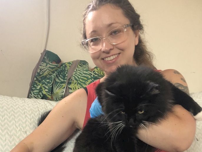 A smiling woman with glasses holds a fluffy black cat on a couch, indoors, with a leafy bag in the background, end this sentence.