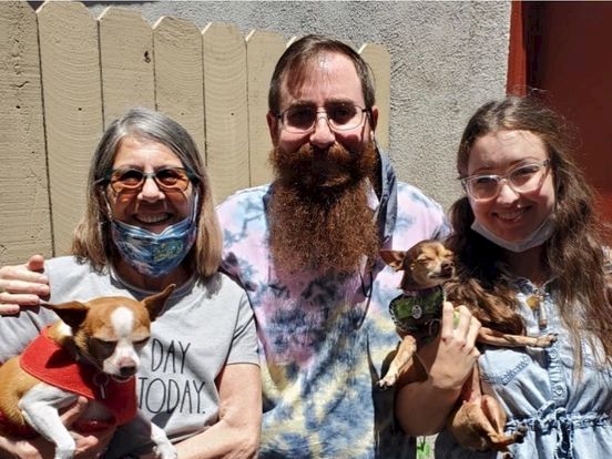 A family with two women and a man, all smiling, holding two small dogs outdoors in front of a wooden fence.