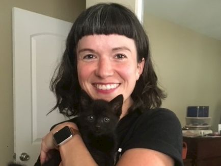 A smiling woman with black hair holding a small black cat; cozy indoor scene in the background, both looking at the camera.
