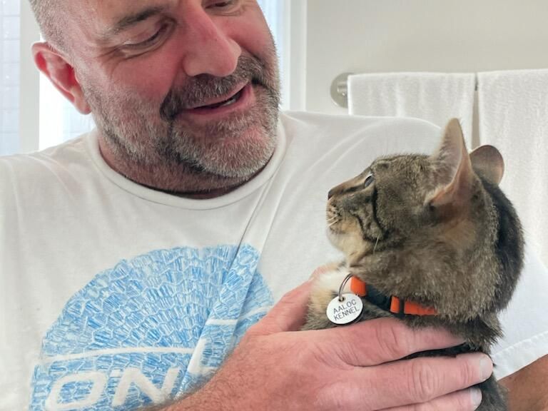 A man with a white t-shirt smiles as he gently holds a striped cat wearing a red collar and tag, sharing a warm moment at home.
