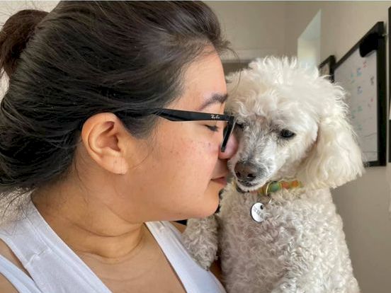 A person in glasses leans in to kiss a fluffy white dog on the nose, both looking at each other lovingly.