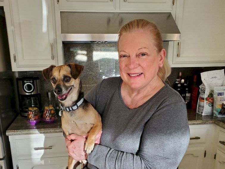 A smiling woman in a gray top holds a small tan dog in a bright kitchen with white cabinets and a stainless range hood.