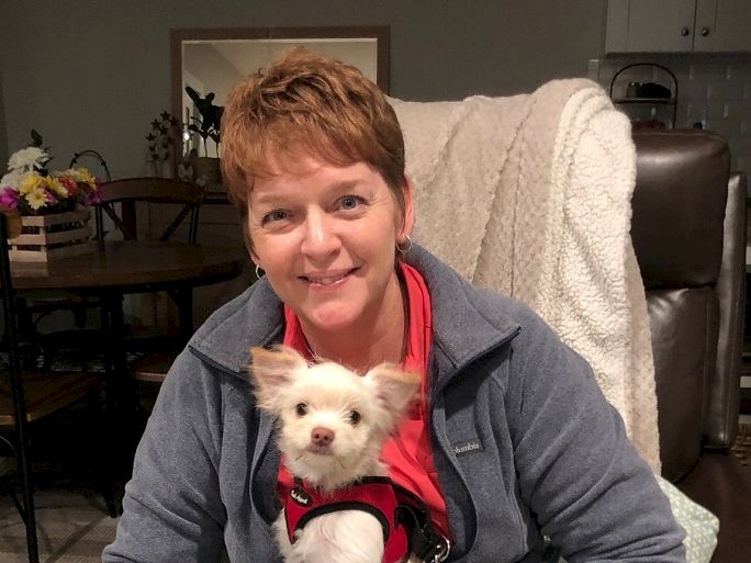 A smiling woman in a gray jacket sits in a chair with a small white dog in a red harness on her lap, cozy home kitchen background.