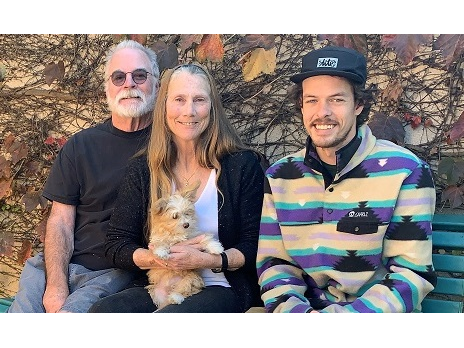 A family of three sits on a bench with a small dog, smiling together outdoors on a sunny day.