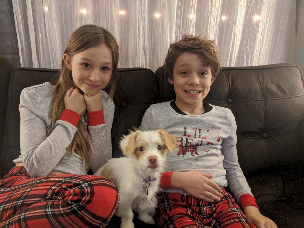 Two kids in matching plaid pajamas sit on a couch with a small white fluffy dog between them, smiling happily at the camera.