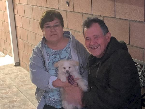 A smiling couple sits on a bench outside a brick building, holding a small fluffy white dog between them.
