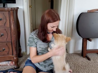 A woman sits on the floor indoors, smiling as she hugs and kisses a small light-colored dog; a dresser and TV stand are in the background.
