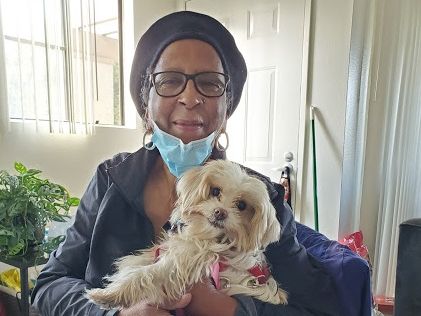 An older woman with glasses and a headscarf holds a small fluffy dog indoors; she has a mask lowered and a gentle smile.
