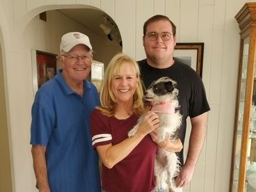 A smiling family of three poses with a small dog indoors, all close together and looking at the camera.