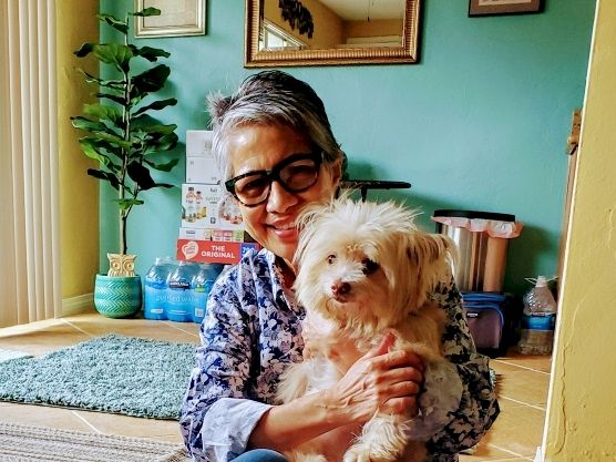 A smiling woman in glasses holds a fluffy white dog indoors, with colorful d&eacute;cor and a plant in the background.