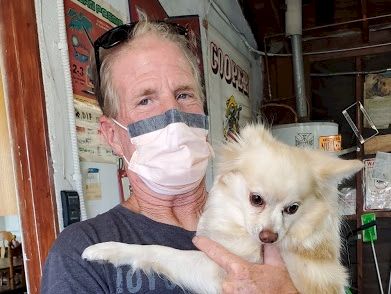 A man wearing a mask holds a fluffy white dog-like creature with one paw raised, against a shop backdrop.
