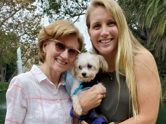 Two smiling women hold a small white dog by a lake, posing for a cheerful outdoor photo.
