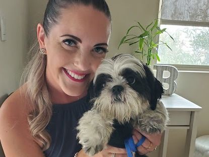 A smiling woman holding a fluffy black-and-white dog, indoors near a window, with a plant and cozy decor in the background.