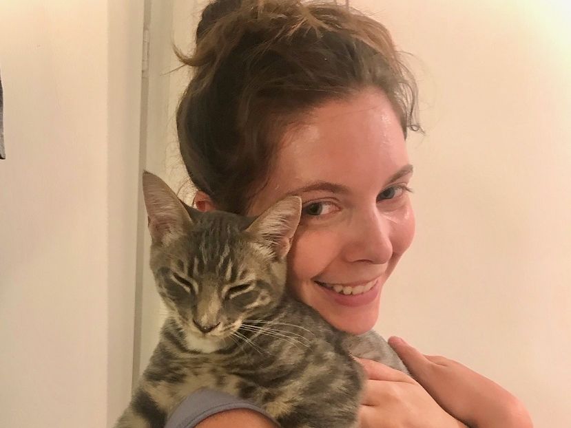 A smiling woman holds a gray tabby kitten, posing for a selfie against a light background. She hugs the cat close and looks happy.