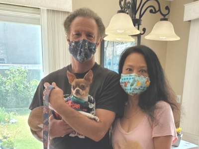 Two people wear masks and hold a small dog; they stand indoors with a window and chandelier in the background.