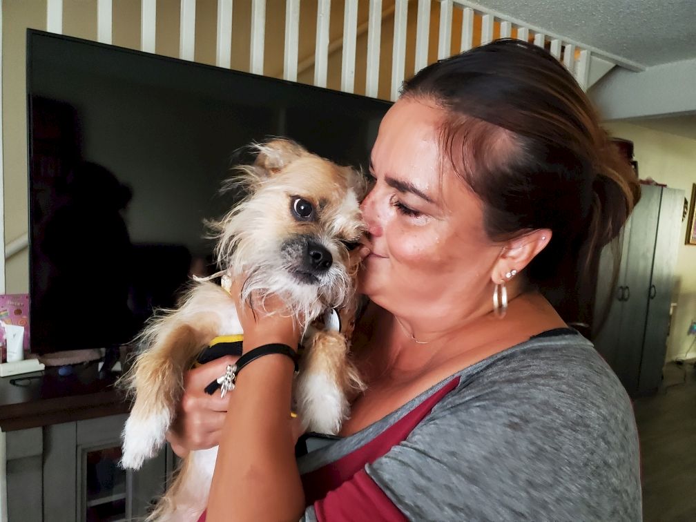 A woman cuddles a small scruffy dog, kissing its face as they share a tender moment indoors.