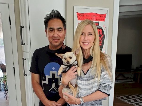 Two people pose indoors with a small dog; the man in black, the woman in a striped sweater, all smiling at the camera.