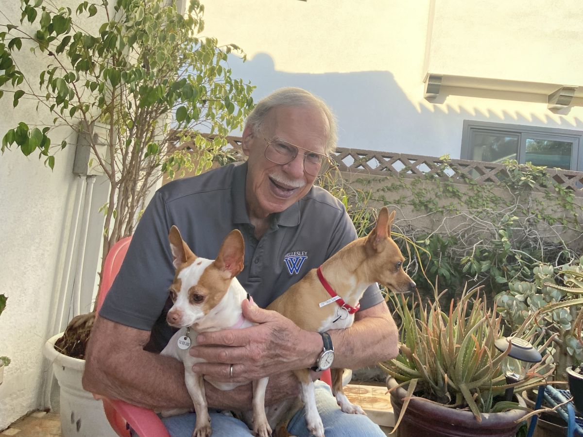 An elderly man sits at a patio, smiling while hugging two small dogs (a white-brown chihuahua and a tan chihuahua) on his lap, in an outdoor garden.