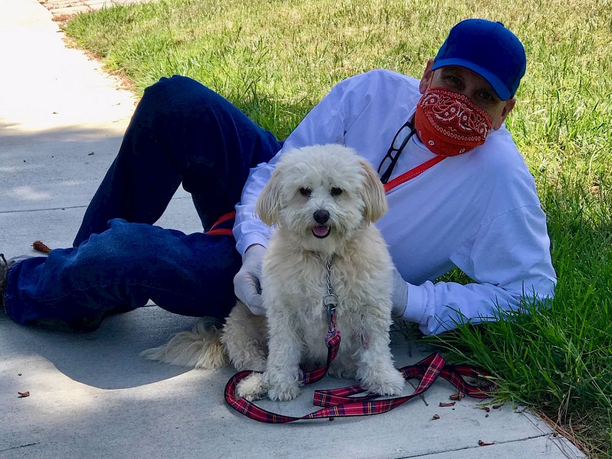 A man in a blue cap and red bandana sits on a sidewalk with a small fluffy white dog on a leash, both outdoors in sunny grass.